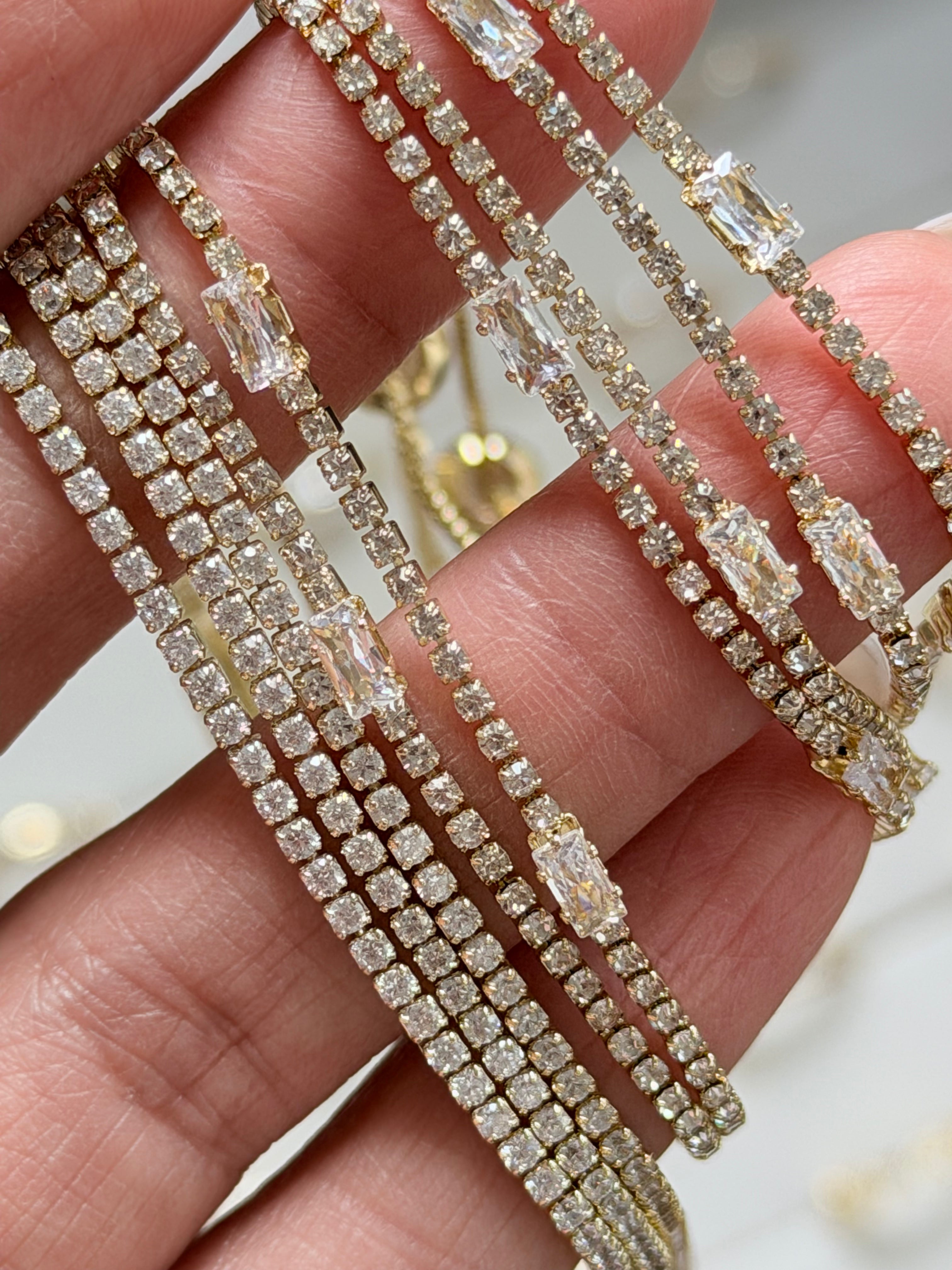 A close-up of a hand holding several gold-toned multi-strand tennis bracelets adorned with clear baguette and round-cut crystals.